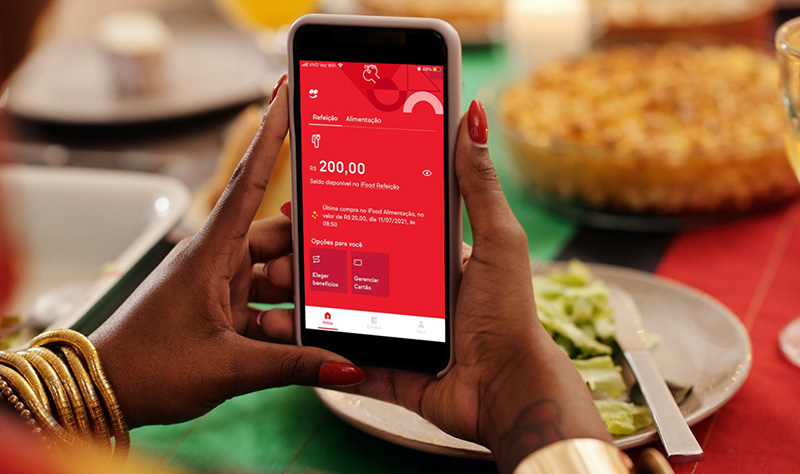 mulher segurando o celular, e na tela o aplicativo do ifood benefícios em fundo vermelho